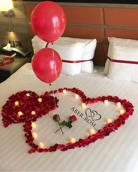 اتاق خواب عروسی که روی روتختی سفید آن گلبرگ های رز و شمع شکل قلب را تشکیل داده اند و شاخه گل ها رز توسط بادکنک های الیومی روی آن قرار داده شده اند